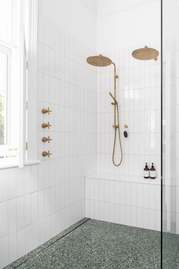 Bright, compact walk-in shower bathroom with white vertically gridded wall tiles, a green-grey terrazzo floor, a built-in tiled bench, and brushed brass shower fittings including dual rain showerheads and a handheld wand. A window provides natural light, while a frameless glass screen is visible at right. The overall look is clean, refined, and contemporary with warm brass accents.