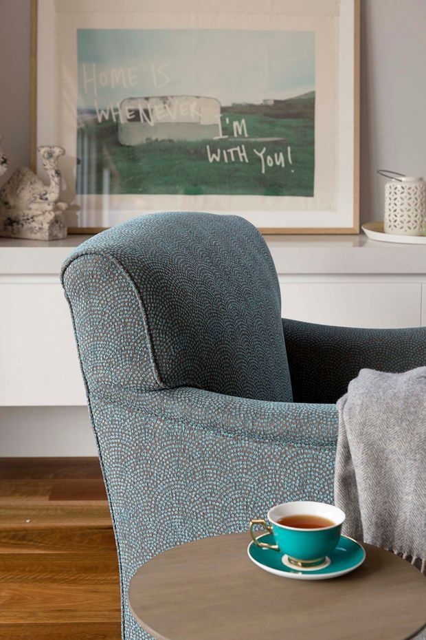 A close-up interior vignette featuring a patterned blue-grey upholstered armchair beside a small round side table with a turquoise teacup and saucer. A light wood floor, white built-in cabinetry or wainscot, framed artwork, decorative objects, and a soft grey throw create a cosy, refined sitting area.