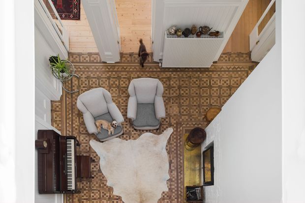 Top-down view of a compact, warmly styled sitting room or entry sitting area. The room features an ornate patterned tile floor, pale timber wall and ceiling finishes, white decorative trim and cabinetry, two upholstered armchairs, a cowhide rug, a dark upright piano, indoor plants, and small decorative objects. The composition is bright and inviting with traditional detailing and a strongly patterned floor.