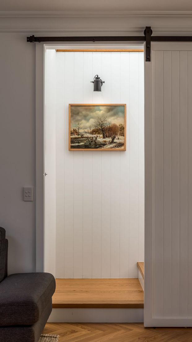 Interior view of a compact transition between a living area and a small raised room or nook, framed by a partially open white vertical-panel sliding barn door with dark metal hardware. The space features warm timber flooring and a timber step, white painted panelled walls, traditional cornice detailing, a small wall light, and framed landscape artwork. A dark upholstered sofa is partially visible in the foreground.