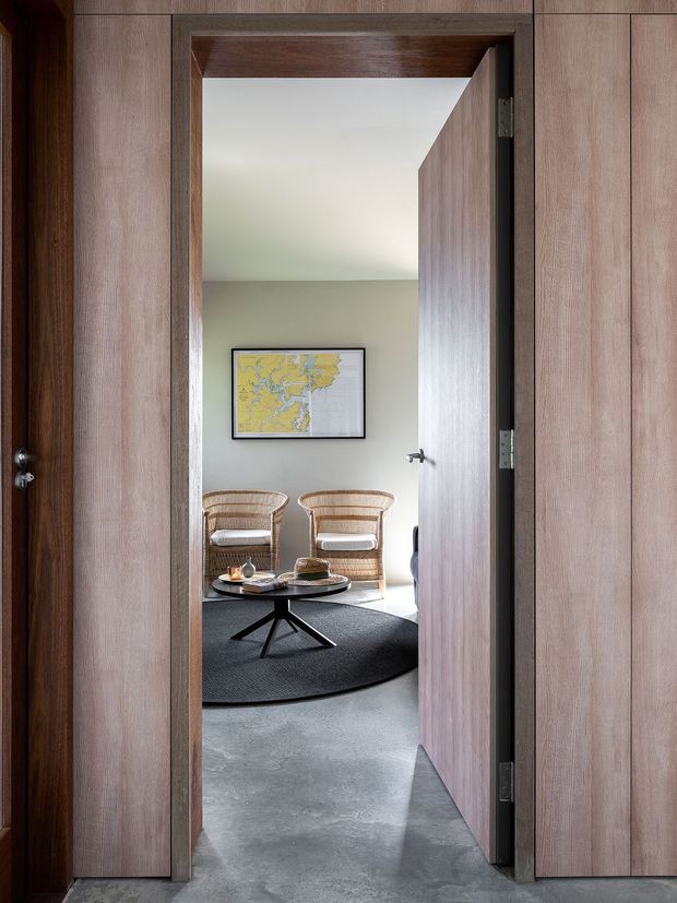 View through a tall timber-lined doorway into a calm sitting area. The room contains two woven lounge chairs, a low black coffee table, a dark round rug, pale walls, a framed map, and a smooth grey concrete floor. The open door and surrounding wall panels are finished in warm medium-to-dark timber.