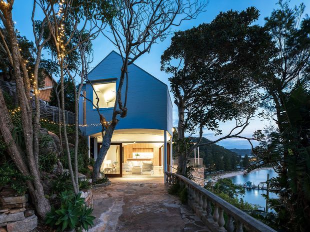 A contemporary multi-level house set on a steep, landscaped waterfront site, viewed along a stone-paved walkway and balustrade. The building features a sharply pitched gabled roof form, blue-grey exterior walls, large illuminated openings, and a warm modern interior visible through the lower glazing. Dense established trees and planting frame views toward the water and neighbouring shoreline.