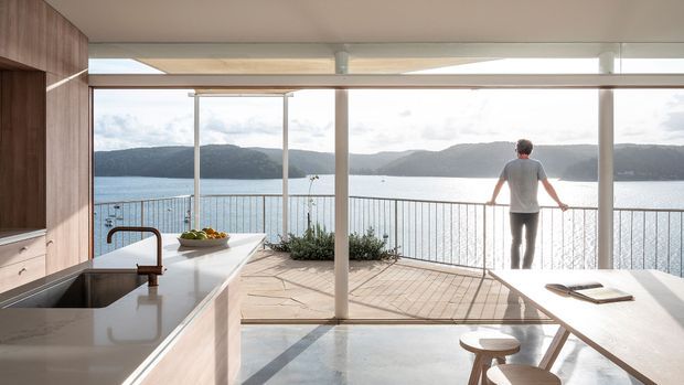 A bright, open-plan kitchen and dining area with warm timber cabinetry, a pale stone island and broad floor-to-ceiling glazing opening onto a balcony or terrace. The space overlooks a calm body of water and wooded hills, with a person standing at the balustrade. Minimal furnishings and strong indoor-outdoor connection create an airy contemporary setting.