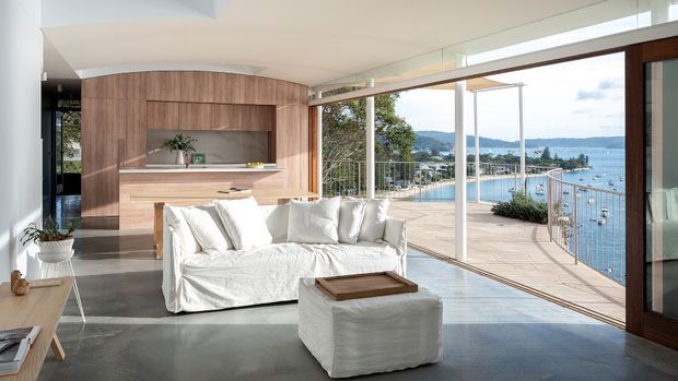 A bright contemporary living room opening onto a large waterfront terrace, with floor-to-ceiling glazing, a curved timber built-in joinery wall, white slipcovered sofa and ottoman, and grey polished flooring. A bay and surrounding hills are visible beyond the terrace.
