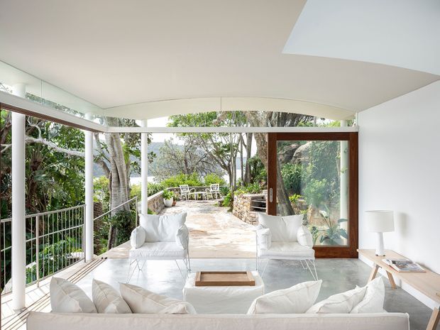 Bright, minimalist living room with a white sofa, armchairs, low coffee table, pale tiled floor, and expansive floor-to-ceiling glazing. A large timber-framed glass door opens onto a stone terrace surrounded by lush vegetation, with a distinctive angular ceiling and white built-in cabinetry.