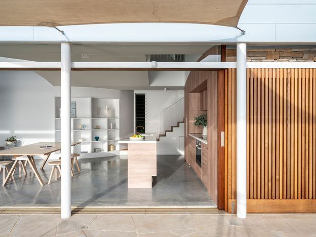 A bright, open-plan contemporary home interior viewed through fully opened glazed doors. The space combines a dining area at left, a compact kitchen with a pale central island, white open shelving, a staircase, and warm timber built-in cabinetry. Polished grey concrete flooring, white structural columns, extensive glazing, and a vertical timber slatted door or screen create a clean indoor-outdoor connection.