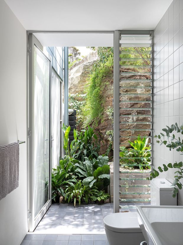 Bright contemporary bathroom with white tiled walls, a freestanding white bathtub and toilet, and a full-height glazed door opening directly onto a lush planted courtyard beside a natural rock face. A vertical louvred window provides ventilation and filtered light, while grey tiled flooring and a wall-mounted towel rail complete the clean, airy fit-out.