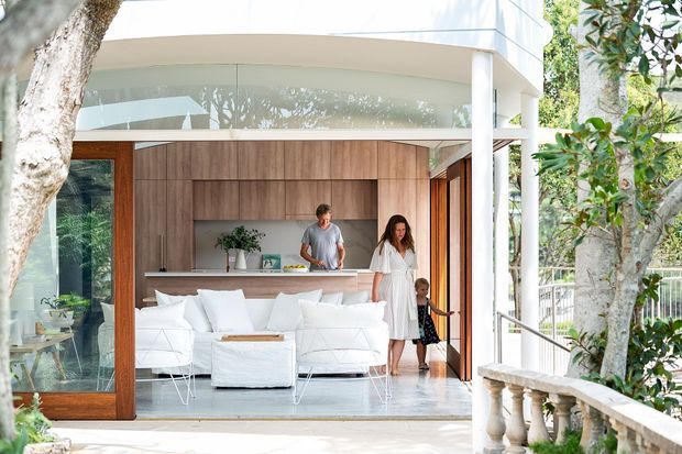 A bright contemporary indoor-outdoor living space with a white lounge area opening onto a terrace, featuring floor-to-ceiling glazing, warm timber-framed doors, a curved white ceiling edge, and a full-height timber feature wall with integrated cabinetry and a kitchen or bar counter. Vegetation and a balustraded exterior terrace surround the space.