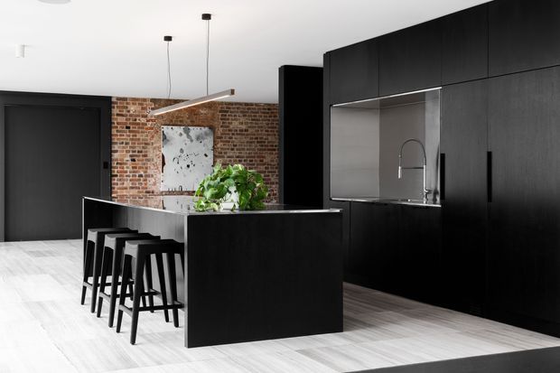 A contemporary open-plan kitchen dominated by a large black island with integrated seating for four. Floor-to-ceiling dark cabinetry forms a streamlined wall of storage and incorporates a stainless-steel sink and curved faucet. A brick feature wall, pale timber flooring, white ceiling, and minimalist pendant lights create a high-contrast, refined industrial-inspired interior.