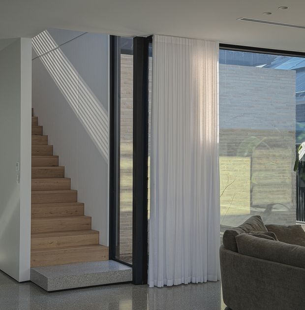 Contemporary interior view of a bright stair hall and living area. A straight timber staircase rises along a white vertically textured wall, beside full-height black-framed glazing and sheer white curtains. The polished grey floor, pale masonry courtyard wall visible outside, and partial curved upholstered sofa create a refined, minimalist composition with strong diagonal sunlight shadows.