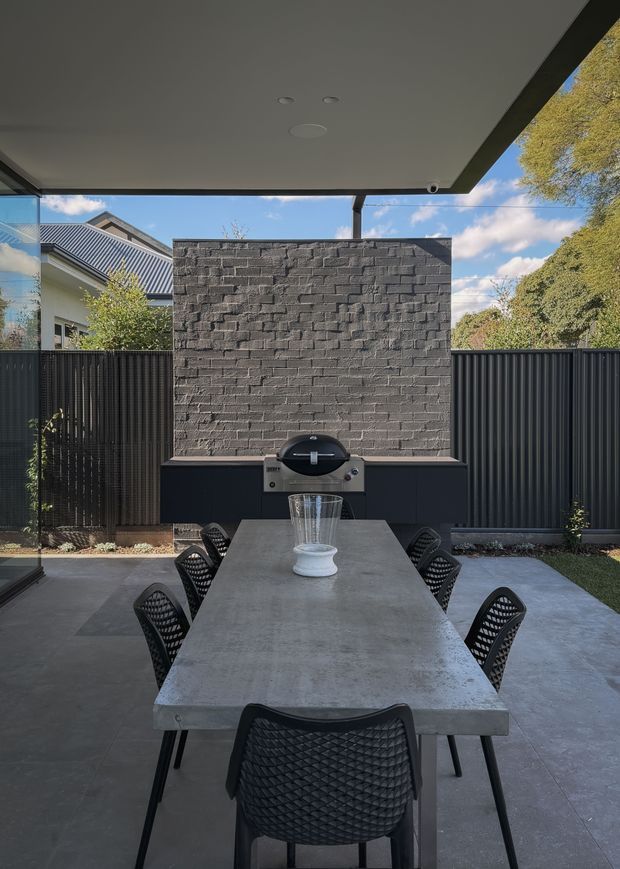Covered outdoor dining area with a long concrete dining table and black woven chairs, opening onto a landscaped courtyard. A built-in barbecue is set against a dark textured brick feature wall, with black metal fencing and glimpses of neighbouring rooflines and trees beyond.