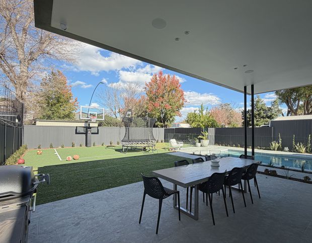 A covered outdoor entertaining area with a dining table and black chairs overlooking a landscaped backyard. The space includes a swimming pool, artificial-turf sports court with basketball hoop and trampoline, dark privacy fencing, and a broad flat roof with recessed downlights supported by slender black posts.
