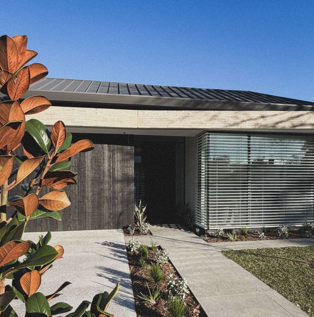A single-storey contemporary residential facade viewed from the front, featuring a low-pitched standing-seam metal roof, pale horizontal masonry cladding, dark timber-look entry doors, and a projecting glazed section with horizontal exterior blinds. A paved path and simple landscaped planting lead to the recessed entrance, with a lawn and clear blue sky visible.