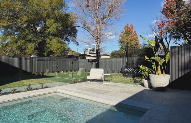 A landscaped residential backyard with a swimming pool in the foreground, surrounded by large-format pale paving, dark vertical-panel fencing, and a glass pool safety barrier. Beyond the pool is a compact artificial-turf sports court with a basketball hoop, lounge seating, potted plants, and mature trees. The scene is sunlit with a clean, contemporary outdoor design.