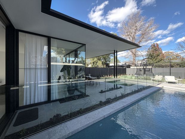 Contemporary residential poolside view with a broad glass-walled living area opening onto a narrow tiled pool. The architecture features a deep flat roof overhang, dark slender framing, floor-to-ceiling glazing, and a minimalist covered outdoor edge. A fenced lawn and outdoor seating are visible beyond the pool.