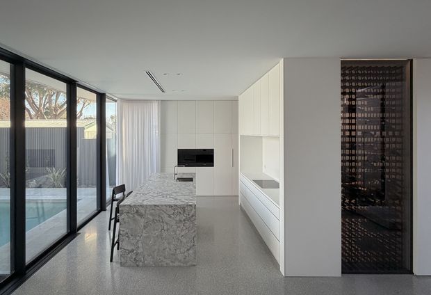 A bright, minimalist contemporary kitchen viewed from the living area, with a large white-and-grey veined stone island, integrated white cabinetry, dark integrated appliances, and a full-height black-framed glazed wall opening to a pool courtyard. A dark patterned feature door or screen is visible at right. The space has a clean, restrained palette and strong indoor-outdoor connection.