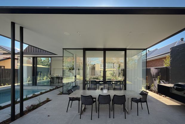 A contemporary covered outdoor dining and entertaining area with a large rectangular table and black chairs, enclosed by extensive floor-to-ceiling glazing. The space connects visually to an interior dining area and a narrow swimming pool, with a neighbouring brick house visible beyond. A broad flat roof, slim dark frames, concrete-look paving and open glass walls create a modern indoor-outdoor connection.