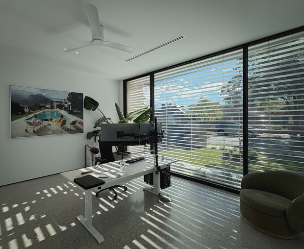 Bright contemporary home office with a height-adjustable white desk, multiple monitors, office equipment, and an ergonomic chair. Full-height black-framed glazing with horizontal blinds occupies one wall, casting striped sunlight across the carpeted floor. A sculptural ceiling fan, indoor plants, and a rounded lounge chair complete the room.