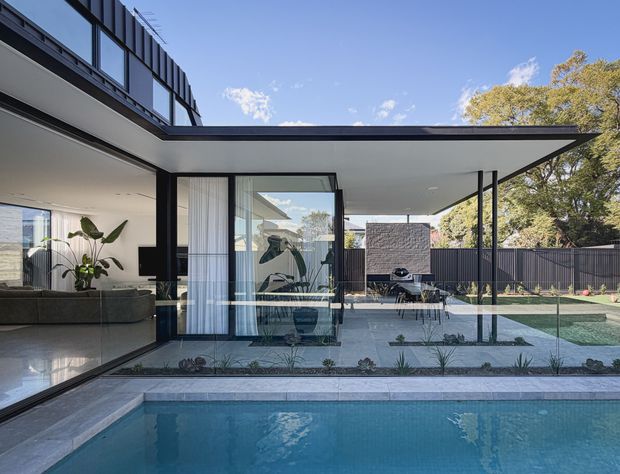 Contemporary single-storey residence with extensive floor-to-ceiling glazing opening onto a covered outdoor entertaining area and swimming pool. The home features a dramatic flat projecting roof, slim dark window frames, polished light flooring, and a dark privacy fence. A living room with a sectional sofa and indoor planting is visible through the glazing, while the sheltered terrace includes outdoor dining and a barbecue area.