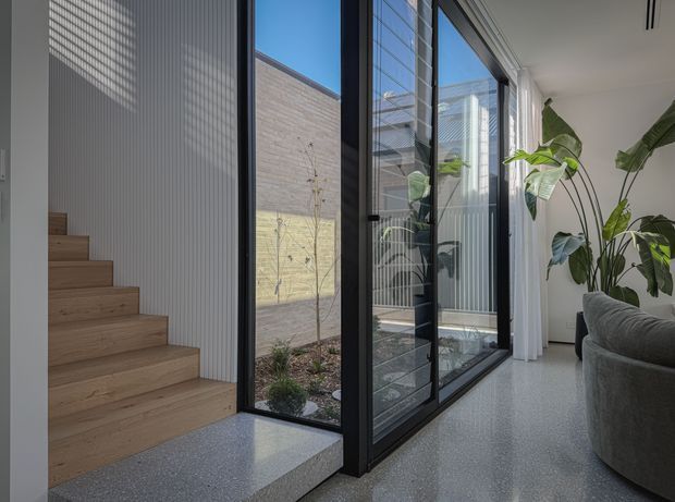 Bright contemporary interior showing a timber staircase beside full-height black-framed glazing opening onto a small landscaped courtyard. The space has polished light terrazzo-style flooring, white vertically ribbed wall panelling, abundant indoor plants, and a partial grey sofa, creating a calm connection between living space and courtyard.