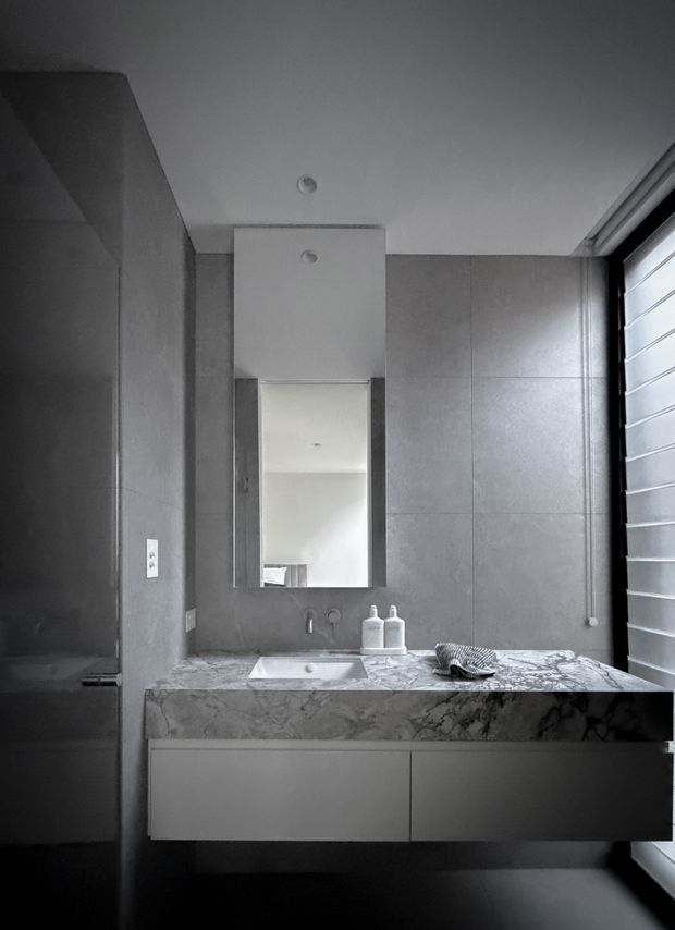A compact, monochromatic contemporary bathroom vanity. The room features large grey wall tiles, a floating white vanity with a heavily veined marble benchtop, integrated basin, wall-mounted tapware, and a rectangular mirror beneath a boxed ceiling bulkhead. A louvred window or shuttered glazing occupies the right side, bringing in natural light. The image has a clean, refined and slightly moody atmosphere.