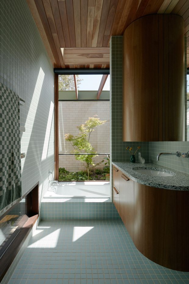A bright, narrow bathroom with a freestanding bathtub positioned beneath a large glazed opening, pale blue-green square tiles covering the floor and walls, and a curved timber vanity with a speckled stone benchtop and integrated basin. The ceiling is lined with warm timber boards, while a patterned tile feature wall and greenery visible beyond the glazing add visual interest. Strong sunlight creates a calm, organic atmosphere.