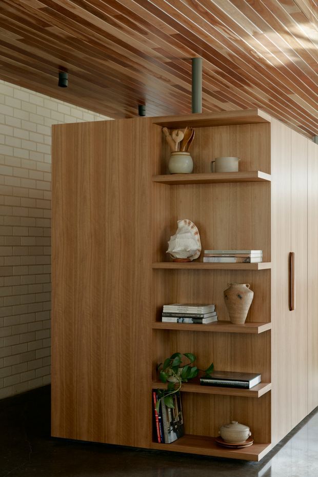 Interior view dominated by a tall, warm timber built-in storage and display unit with open shelving, concealed cabinetry, and a vertical wood handle. The joinery stands against a pale brick-tile wall and beneath a richly timber-lined ceiling with recessed black spotlights and a dark structural post. Shelves display books, ceramics, sculptural objects, and a small plant. A polished dark floor and bright adjacent circulation area are visible at the base and right edge. The composition has a refined, warm, natural contemporary character.