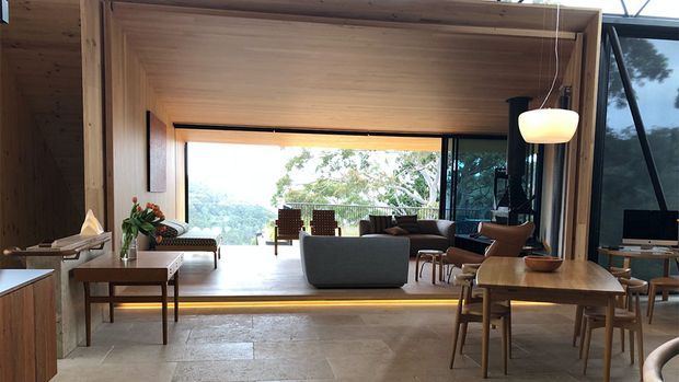 Open-plan contemporary living room with a timber-lined portal framing a raised sitting area. The room features full-height glazing, warm wood surfaces, pale stone flooring, mid-century-inspired lounge furniture, and a sculptural pendant light, with a leafy landscape visible outside.