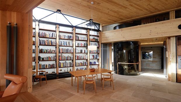 A warm, expansive open-plan living and dining space with extensive timber detailing, a large built-in library wall, a timber dining table and chairs, and a double-height glazed area. A mezzanine-like timber bridge spans the room above a central fireplace or hearth, while stone or tile flooring and exposed timber ceilings create a refined rustic atmosphere.
