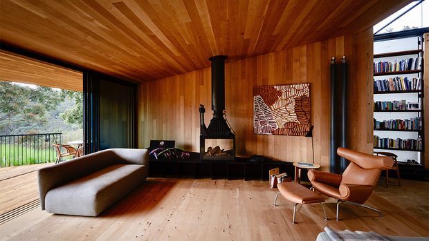 Warm, timber-lined living room with a full-height wood ceiling and walls, pale timber flooring, a sculptural black wood-burning fireplace, built-in shelving filled with books, and mid-century lounge furniture. Large glazed openings connect the room to a leafy outdoor setting and deck.