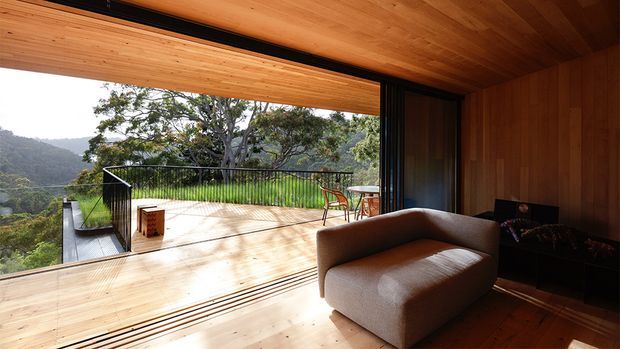 A warm timber-lined living room opens through fully retractable glazing onto a spacious elevated deck. The room features a low contemporary sofa, dark built-in cabinetry, timber flooring and ceiling, and expansive views over a lush mountainous landscape. The deck has a slim black balustrade, outdoor dining furniture, and integrated bench seating.