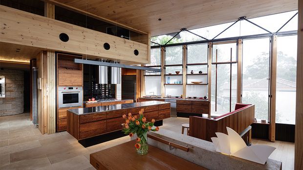 Open-plan contemporary kitchen and living space with extensive timber detailing, a large central island, integrated stainless-steel appliances, open shelving, and floor-to-ceiling glazed walls. A raised timber mezzanine or upper-level structure features circular openings, while a built-in timber seating area and dining table occupy the foreground. Natural light fills the warm, material-rich interior.