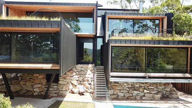 Exterior view of a contemporary multi-level dwelling integrated into a wooded, sloping site. The building features dark vertical metal cladding, extensive glazing, timber-lined upper-level soffits and balconies, exposed steel supports, and substantial natural stone retaining and base walls. A central exterior stair connects the split levels, with a narrow strip of pool water visible at the lower right.