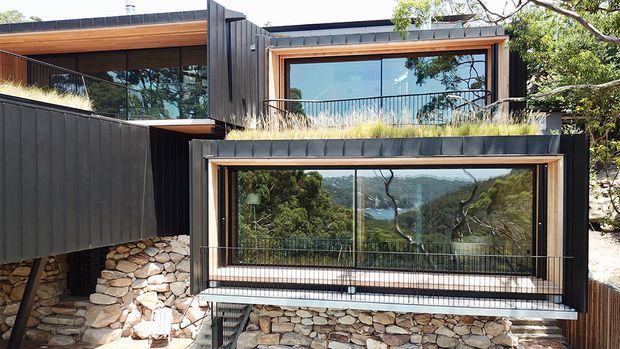 Exterior view of a contemporary multi-level home built into a sloped, wooded site. The facade combines dark vertical metal cladding, extensive glazing with timber-toned window surrounds, planted roof sections, and substantial natural stone retaining walls. A projecting glazed volume and balconies overlook a green valley or coastal landscape.