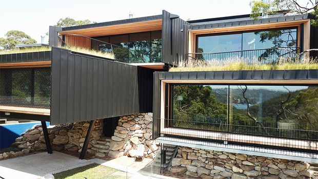 Modern multi-level house integrated into a sloped, rocky landscape, with dark vertical cladding, extensive floor-to-ceiling glazing, warm timber soffits, planted roof edges, and stone retaining walls. The elevated structure overlooks a wooded valley or coastal landscape.