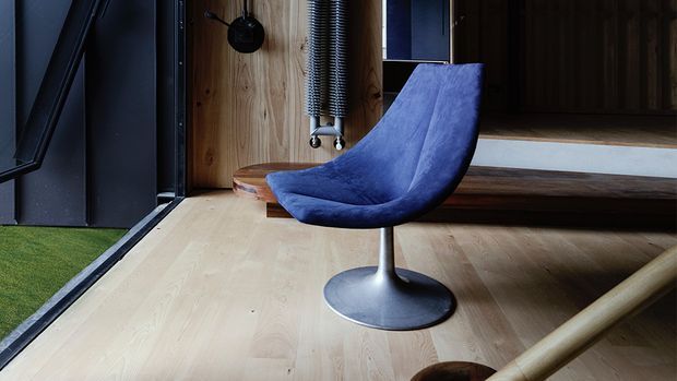 A moody contemporary interior corner featuring a sculptural deep-blue lounge chair on pale timber flooring. Dark metal-framed glazing, a vertical timber wall with exposed mechanical detailing, and a raised timber platform or bench create an industrial architectural setting.