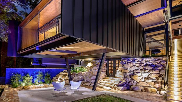 A dramatic modern multi-level home exterior at night, featuring cantilevered box-like volumes with dark vertical cladding, warm timber soffits, extensive glazing, exposed angled supports, and a naturally stacked stone retaining wall. A landscaped outdoor seating area with sculptural chairs sits beneath the cantilever, illuminated by vivid blue and warm architectural lighting.