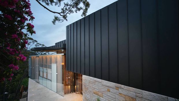Contemporary residential exterior at dusk, featuring a strong black vertically panelled facade above a light stacked-stone wall. Floor-to-ceiling glazed elements and a projecting canopy reveal a warmly lit interior, while surrounding vegetation frames the building on a sloping site.