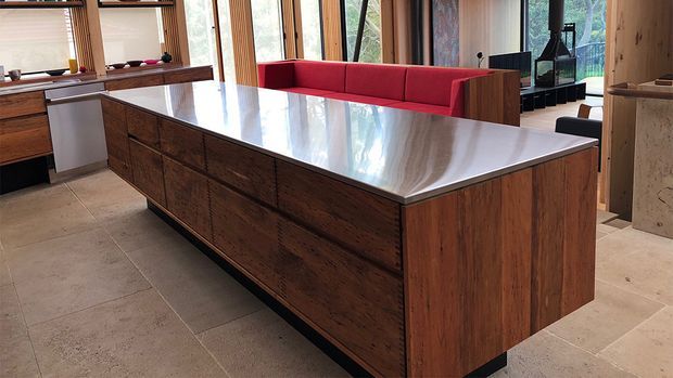 A spacious contemporary kitchen centred on a large island with dark timber cabinetry and a polished light stone benchtop. Tall timber-framed glazing, warm stone flooring, a red upholstered seating area, and built-in timber shelving are visible.