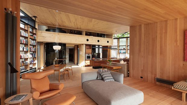 Warm, timber-lined open-plan living room with extensive built-in bookshelves, a large grey chaise sofa, leather lounge chairs, a dining area, and a raised timber mezzanine or bridge with circular openings. Large windows bring in natural light, while the continuous timber floor, walls, and ceiling create a cohesive contemporary interior.