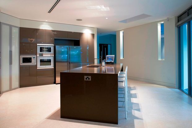 A sleek contemporary kitchen with a large dark glossy island, integrated stainless-steel appliances, tall brown cabinetry, pale flooring, and bar seating. Bright blue daylight enters through full-height glazing and narrow vertical windows, creating a clean, high-contrast interior.