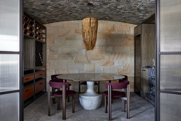 A compact, richly textured dining area enclosed by dark-framed translucent glass doors. The room features a round glass dining table with sculptural pedestal base, burgundy upholstered dining chairs, dark built-in wine storage and cabinetry, a natural stone feature wall, and a dramatic woven pendant beneath a patterned stone-clad ceiling.