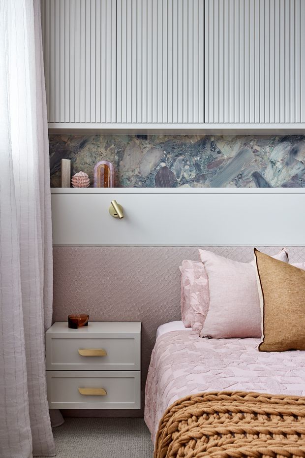 A softly styled bedroom corner featuring a partially visible bed with layered blush pink bedding, pink and tan pillows, and a chunky tan knitted throw. A pale grey bedside table with brass handles sits beside the bed, while a pleated vertical-panel wall, decorative stone-look feature strip, and concealed shelving create a refined built-in backdrop. Sheer curtains frame the left side, and a muted textured carpet covers the floor.