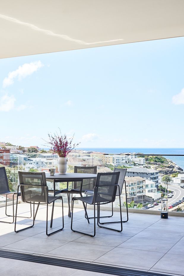 Bright, contemporary dining area opening onto a spacious glazed balcony or terrace, with a dark dining table and mesh chairs positioned against panoramic coastal and urban views. Large frameless glass balustrades and pale tiled flooring create an airy, elevated outdoor-facing setting.