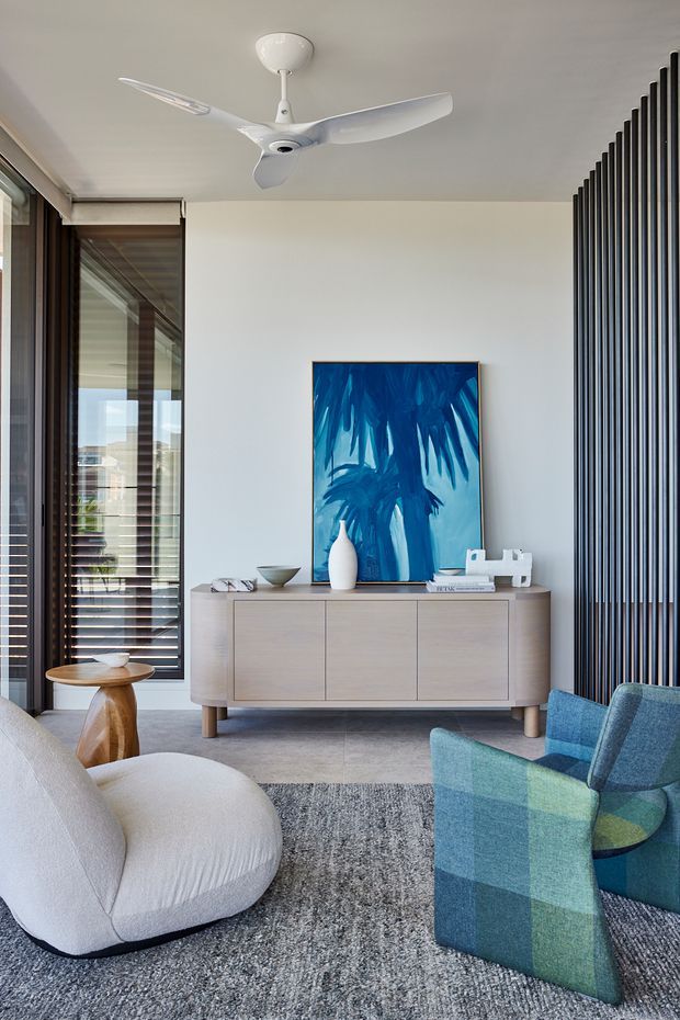 A contemporary living room with a white ceiling fan, pale neutral walls, full-height windows with dark timber or metal framing, and a vertical black slatted feature screen. A light timber sideboard sits beneath a vivid blue abstract artwork, complemented by a sculptural white lounge chair, blue-green armchairs, a small timber side table, and a textured grey rug over a hard grey floor.