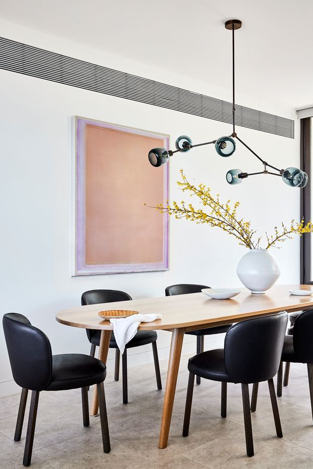Bright contemporary dining room with a long light timber dining table, black upholstered dining chairs, pale tiled flooring, white walls, and a sculptural multi-arm pendant light. A large muted blush artwork and a white vase with yellow flowering branches add restrained colour accents. A linear ceiling air vent and dark-framed glazing are visible.