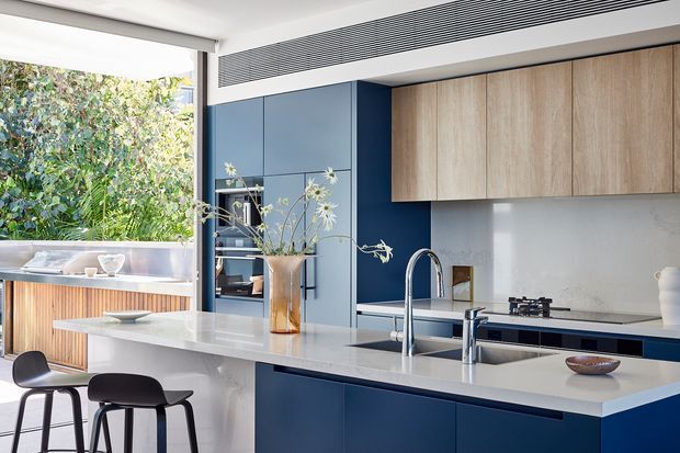 A bright, contemporary kitchen with a large white island and integrated sink, deep navy-blue cabinetry, pale timber overhead cupboards, and a dark stone or composite benchtop. Full-height glazing opens toward lush greenery, while black stools, integrated appliances, and a linear ceiling vent complete the refined interior.