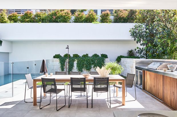 Bright contemporary outdoor entertaining area with a timber dining table and black-framed chairs on a tiled terrace beside a swimming pool. A built-in outdoor kitchen with timber-slatted cabinetry and a stone benchtop occupies the right side, while white rendered walls, glass pool fencing, greenery, and a neighboring building form the backdrop.