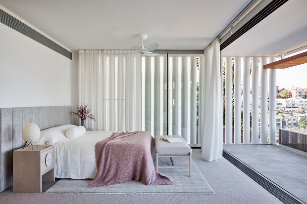 Bright contemporary bedroom with a large upholstered bed, soft grey carpet, sheer white curtains, and floor-to-ceiling glazing opening onto a covered balcony or terrace. Vertical privacy screens, integrated linear air vents, and a ceiling fan contribute to the clean architectural design, while pale timber bedside joinery and muted mauve textiles add warmth.