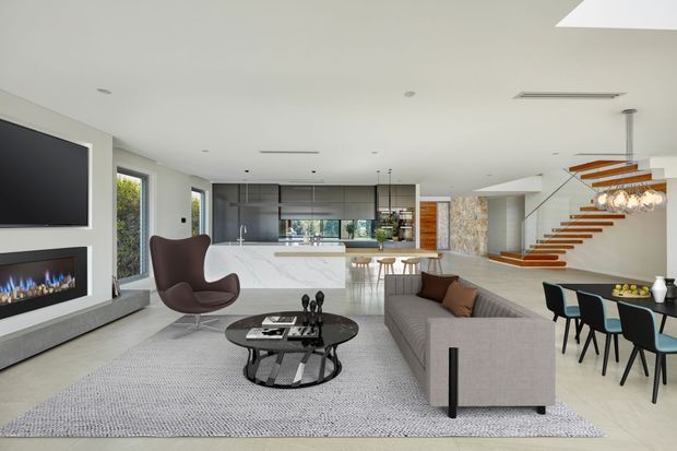 Open-plan contemporary living area with a sleek kitchen, dining zone, and floating staircase. The room features a large linear fireplace and television wall, pale flooring, a grey sectional sofa, sculptural coffee table, designer lounge chair, and a marble-look kitchen island. A stone-clad wall and timber stair treads add textural contrast.